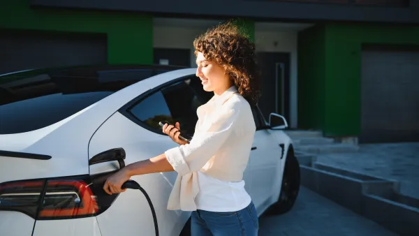 Woman in white shirt charging white electric sedan in front of house.