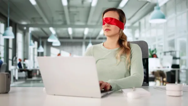 Woman wearing red blindfold sitting at white table typing on silver laptop.
