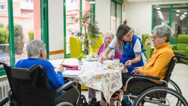 Three senior women sitting at table with caretaker leaning over them in bright group home.