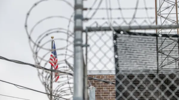 Close-up in barbed wire fence around immigration detention center with American flag visible in background.