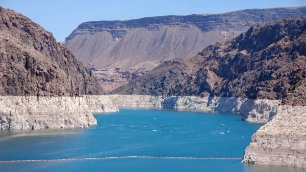 The Hoover Dam blocking the Colorado River. The river's water level is noticeably low. The cliffs surrounding the blue water are brown and jagged.