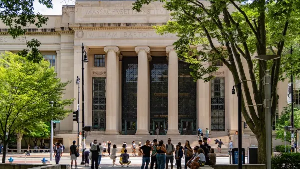 People walk in front of an MIT building with marble pillars. There are trees in the foreground.