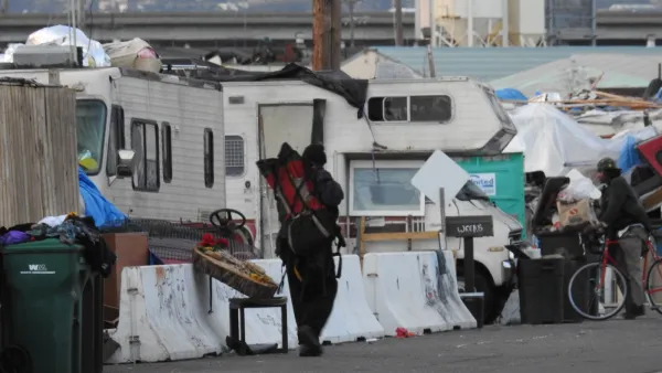 Man walks past homeless encampment behind concrete Jersey barriers on street in Oakland, California with RVs parked behind tents.