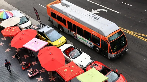 A Metro bus drives down a street on a rainy day. Parallel to the bus is a line of parked cars, then, a line of umbrellas. 