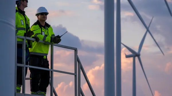 Two construction workers on a wind turbine, with two more in the background against a pink sky.