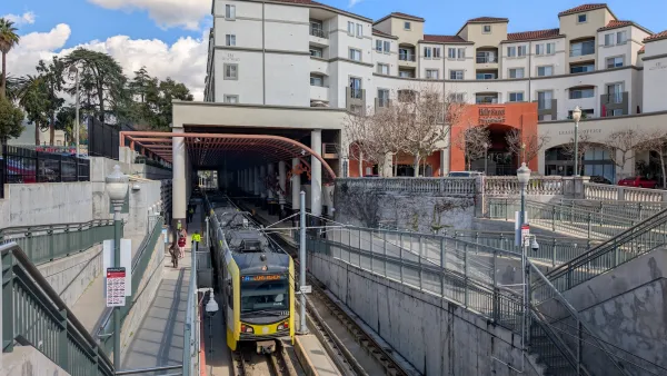 Yellow LA Metro train pulling out of station in Pasadena, California with neo-Spanish style apartment building visible above station.