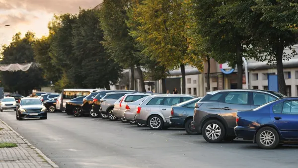 A row of cars parked next to a building. One car drives perpendicular to the parked cars.