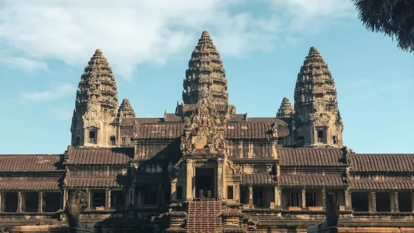 The Ancient Angkor Wat temple in Cambodia captured in early morning light. The temple is gray, and has three pillars with cone shaped tops. It is under a light blue sky.