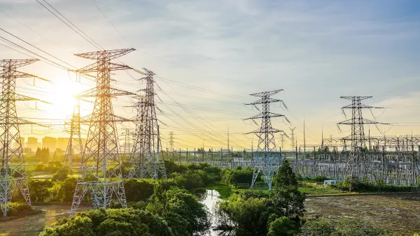 Many large power transmission towers loom over a small river with green trees on either side at sunset.