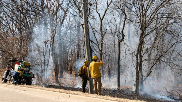 Two firefighters stand at edge of paved road looking at burned, smoldering forest after fire.