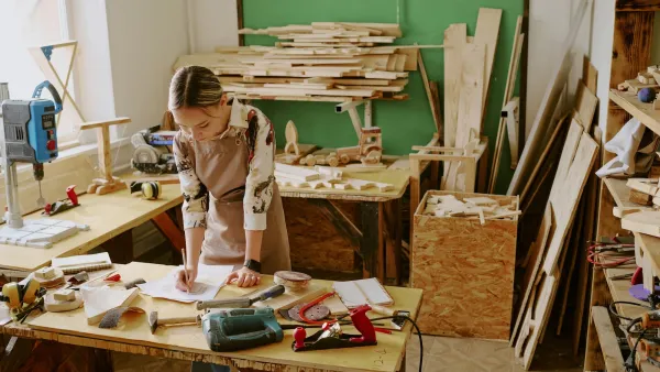 Woman leaning over work table in woodworking space surrounded by lumber.