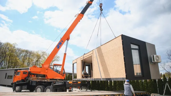 A crane lifts a manufactured house, with is rectangular and does not yet have a roof.