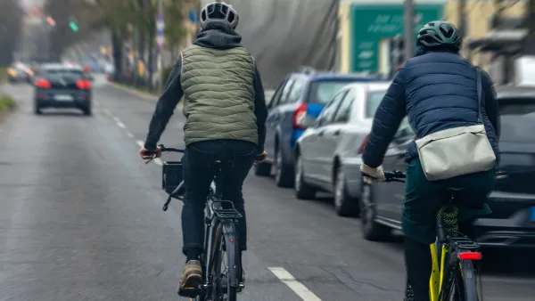Two people on bikes in puffer jackets riding on street next to row of parked cars.