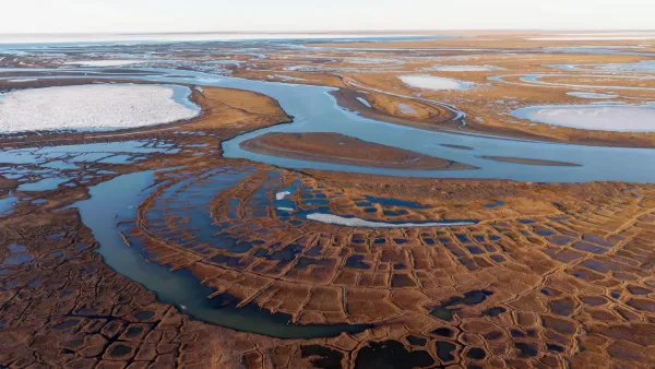 An aerial view of Alaska's National Petroleum Reserve. Patches of residual snow and ice dot the tawny tundra wetlands stretching toward a frozen lake on the horizon. 