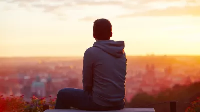 View from back of young man sitting on concrete bench overlooking blurred city at sunrise or sunset