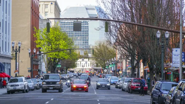 A road in Portland with cars facing the camera and tall buildings on either side. The cars are stopped at a traffic light.