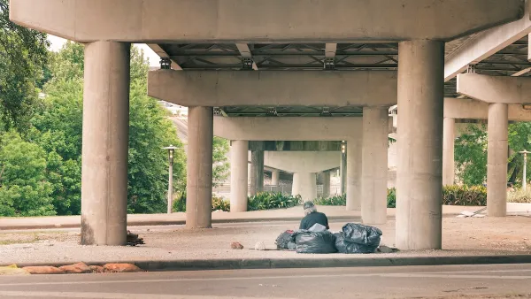 Man with grey hair sits next to black garbage bags in freeway underpass in Houston.