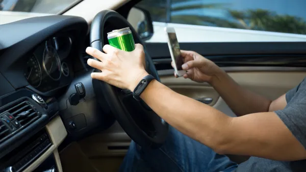 Close-up of man's arms in driver's seat with one hand holding green can on steering wheel and other hand holding cell phone.