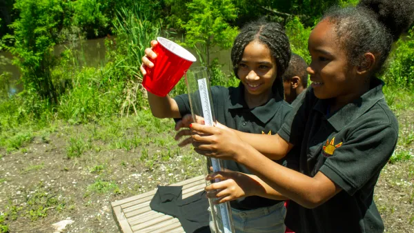 Two young Black girls outdoors using red plastic cup to pour water into tube for measurement or testing.